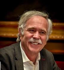 Retrato de un hombre con cabello canoso y bigote sonriendo en un entorno político.