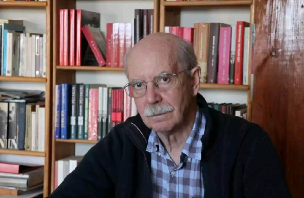 Retrato de un hombre mayor con gafas y camisa a cuadros en una biblioteca.