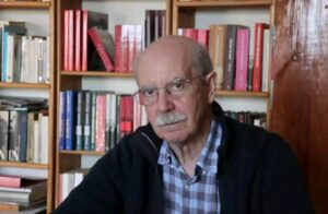 Retrato de un hombre mayor con gafas y camisa a cuadros en una biblioteca.